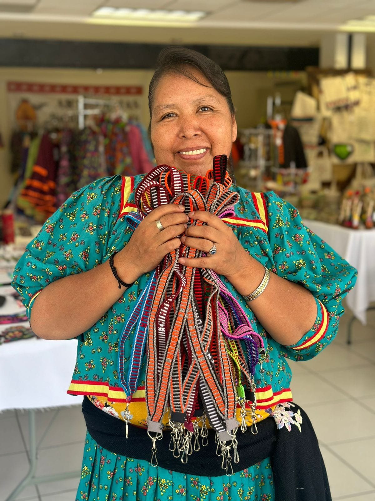 Artesana mostrando varias fajas tejidas con diseños geométricos en rojo, negro, naranja y otros colores.