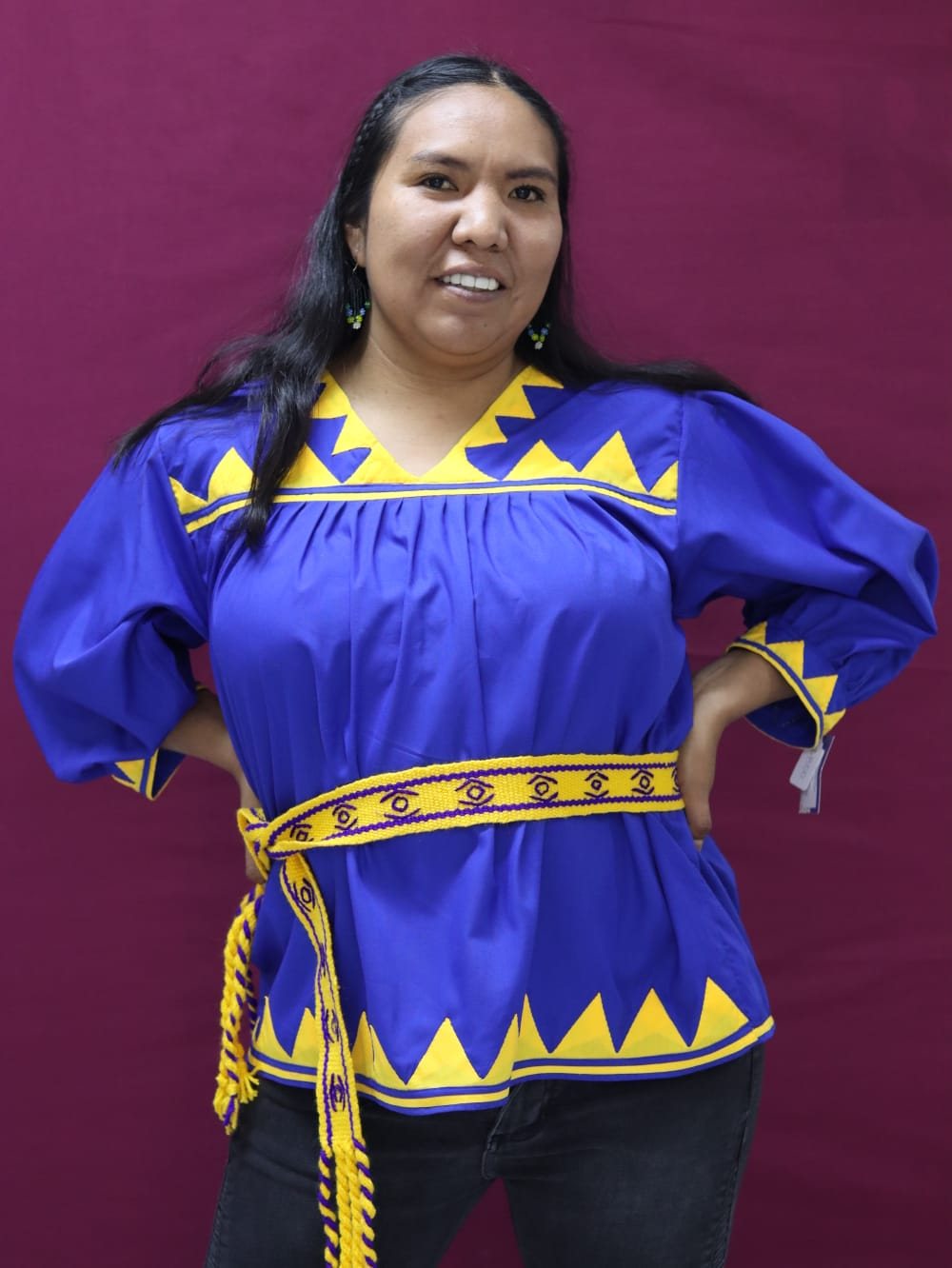 Mujer con blusa morada de mangas amplias, detalles amarillos y faja tejida.