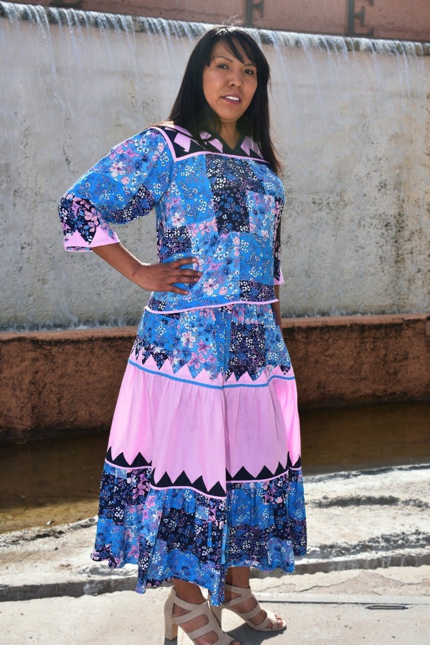 Mujer con blusa azul floral y falda tradicional en tonos azul y rosa.