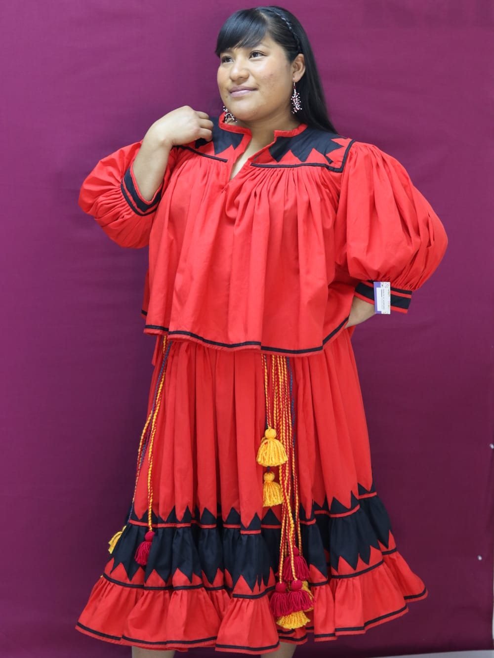 Mujer con conjunto tradicional rojo de blusa amplia y falda, decorado con aplicaciones geométricas negras.