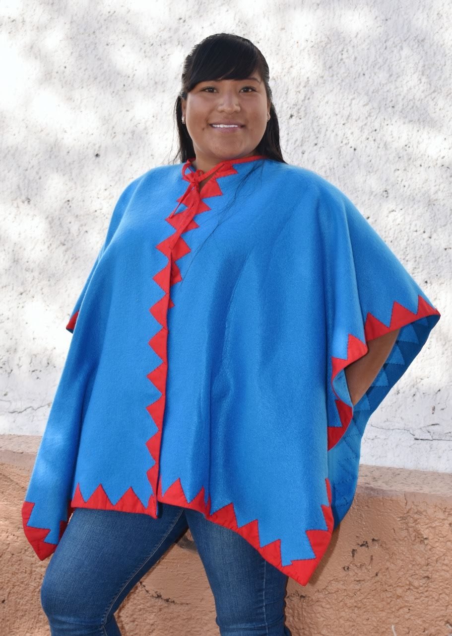 Mujer con poncho azul turquesa y aplicaciones geométricas rojas.