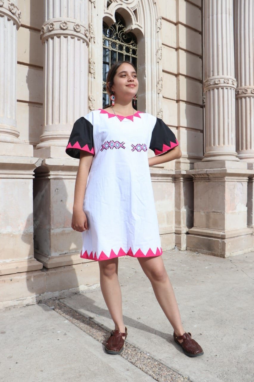 Mujer modelando un vestido blanco con aplicaciones geométricas negras y rosas de inspiración rarámuri.