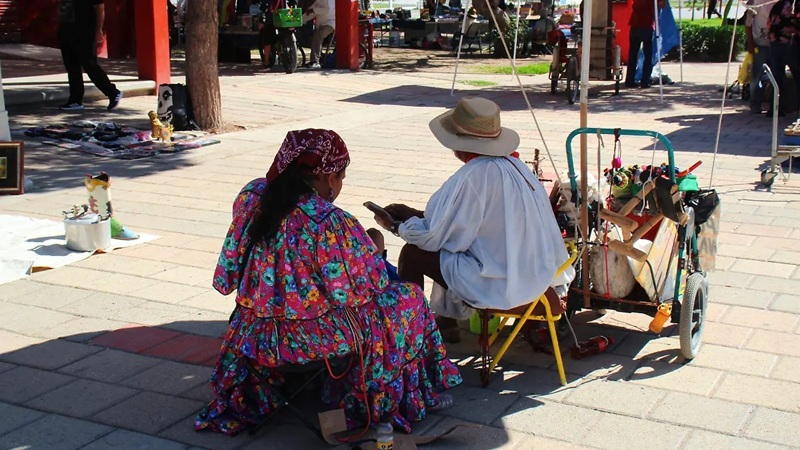 Mujer y hombre de la comunidad Ódami del norte sentados en una plaza pública con vestimenta tradicional y herramientas de trabajo artesanal.