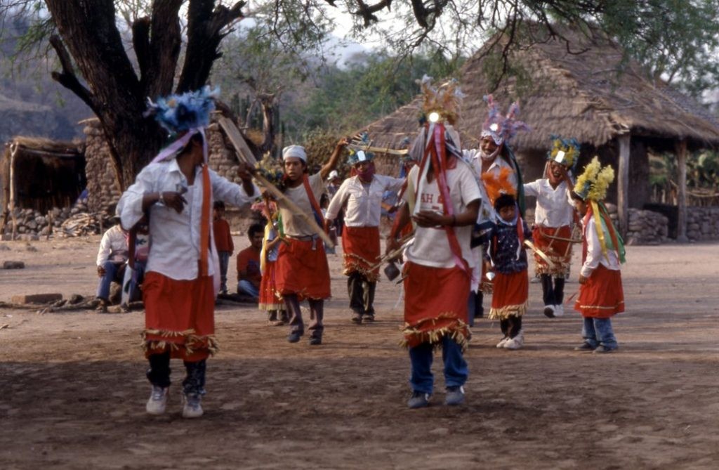 Integrantes de la comunidad Ódami del norte realizando una danza tradicional ceremonial con vestimenta colorida frente a viviendas rurales.