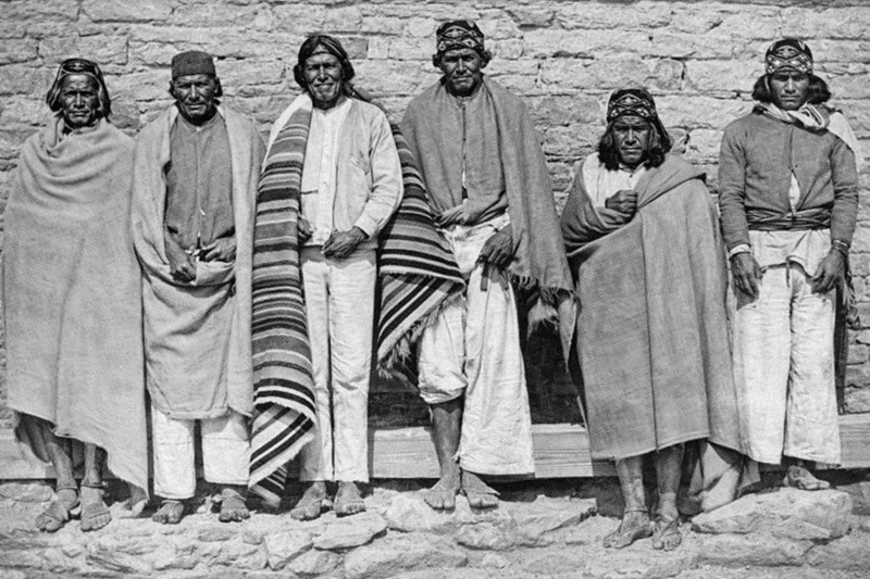 Retrato histórico en blanco y negro de integrantes de la cultura Ódami del norte, de pie frente a un muro de piedra, vistiendo ropa tradicional y cobijas tejidas.e