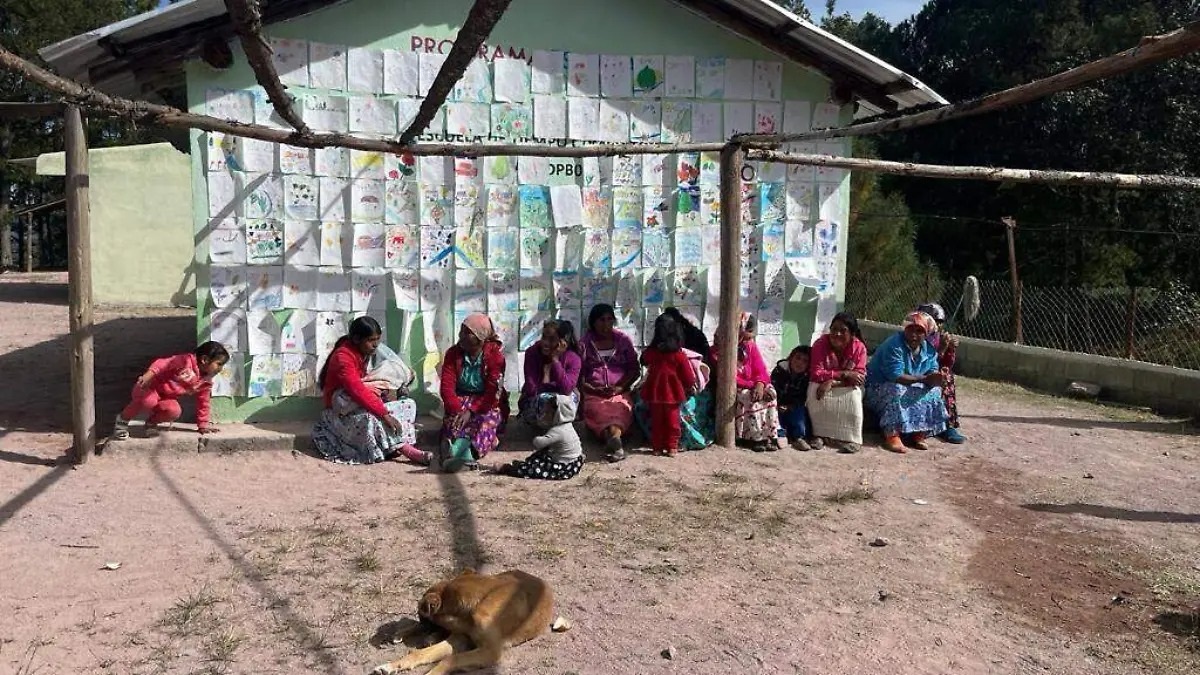 Mujeres y niñas de la comunidad Ódami del norte reunidas frente a una escuela rural con dibujos escolares expuestos en la pared.