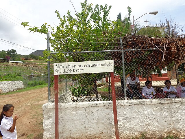 Letrero en lengua Ódami del norte colocado frente a una escuela rural, con niños y paisaje comunitario al fondo.